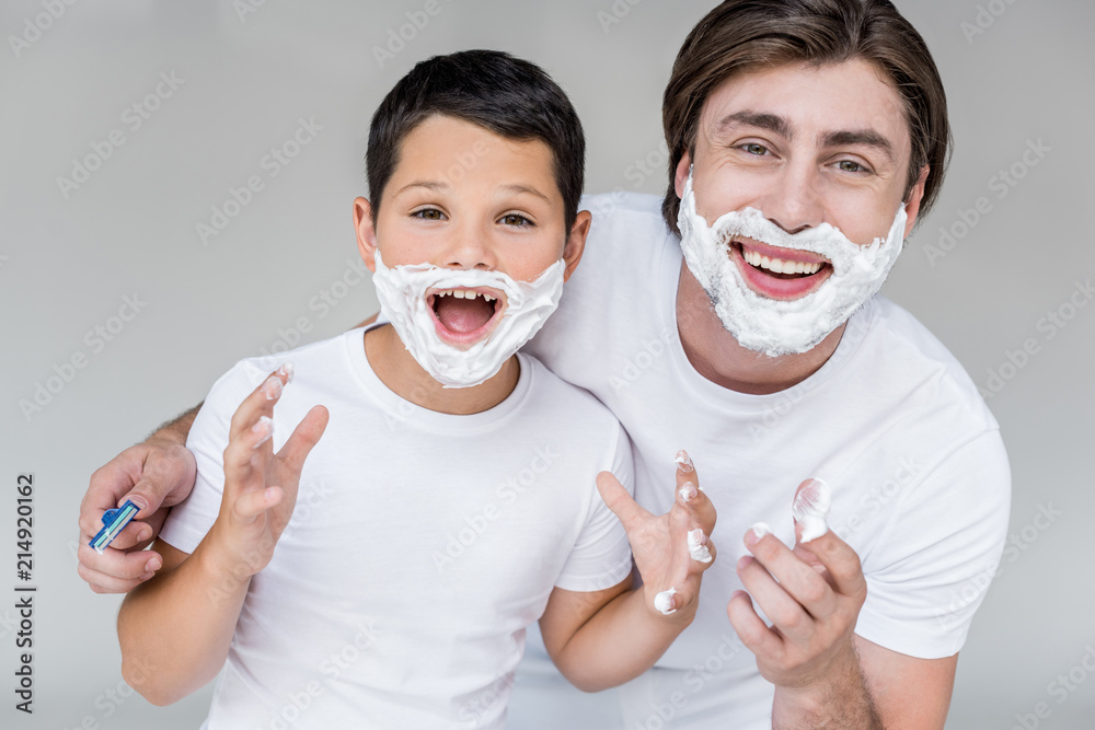 portrait of emotional father and son with shaving foam on faces isolated on grey