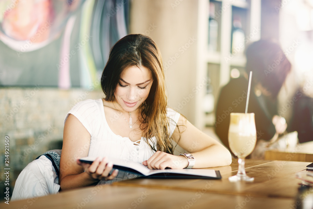 Portrait of a young, beautiful woman sitting alone in a cafe, reading, looking interested and amused; without phone or laptop. Portrait of a young, beautiful woman sitting alone in a cafe, reading, looking interested and amused; without phone or laptop.