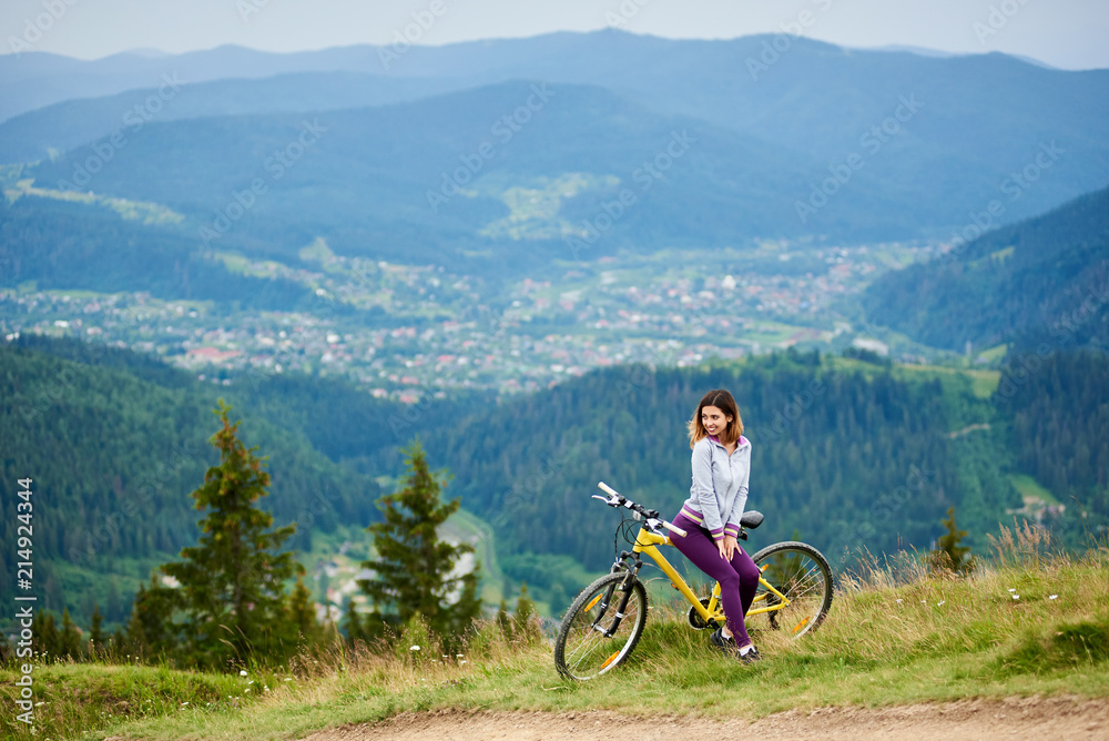 Obraz premium Young woman biker sitting on yellow bike near rural trail on the top of the mountain on cloudy evening. Mountains, forests and small city on the blurred background. Outdoor sport activity