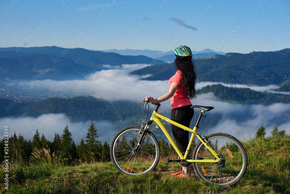 Back view of sexy woman cyclist with yellow bicycle on a rural trail in the mountains, wearing helmet and red red t-shirt, enjoying morning haze in valley, forests on the blurred background