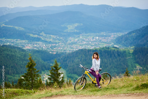 Young woman biker sitting on yellow bike near rural trail on the top of the mountain on cloudy evening. Mountains, forests and small city on the blurred background. Outdoor sport activity