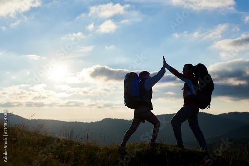 Silhouette of happy couple with backpacks on top of the mountain give each other a high five against the background of the mountains and the cloudy sky with a bright sun at sunset. Bottom view