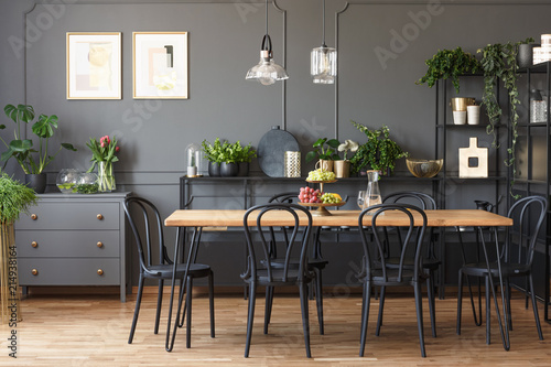 Real photo of a gray and black dining room interior with posters on a dark wall with molding  lamps above wooden table and plants on metal racks