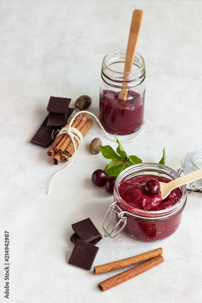 Fototapeta premium Homemade cherry jam with chocolate, spices (cinnamon, nutmeg) with fresh cherries in a jar on a light background.