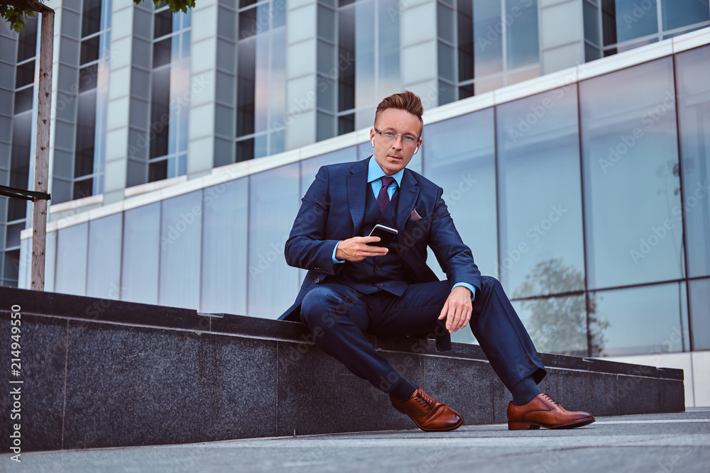 Portrait of a confident stylish businessman dressed in an elegant suit holds a smartphone and looking at camera while sitting outdoors against a skyscraper background. Portrait of a confident stylish businessman dressed in an elegant suit holds a smartphone and looking at camera while sitting outdoors against a skyscraper background.