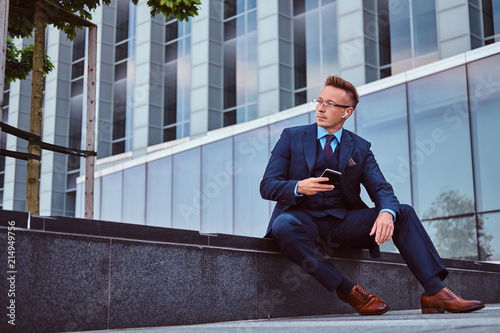Portrait of a confident stylish businessman dressed in an elegant suit holds a smartphone and looking away while sitting outdoors against a skyscraper background.