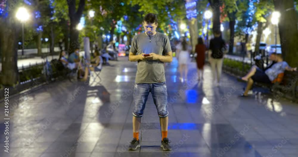 Timelapse of man standing still with smartphone on crowded walkway park boulevard with christmas garlands at night. People moving fast. Concept of flowing time and technologies