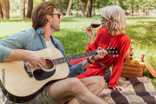 handsome boyfriend playing acoustic guitar for girlfriend at picnic in park
