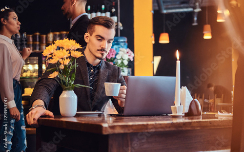 Handsome freelancer man with stylish beard and hair dressed in a black suit working on laptop while sitting at a cafe.