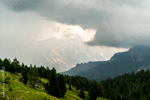  Italian Dolomites landscape. Light after rain in Dolomites. Rocky peaks in the background surrounded by rain clouds. Layers of forest and mountains ridge. Rocky Mountains Dolomiti. Storm rain over r