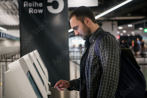Young man with backpack touching interactive display using self service machine, doing self-check-in for flight or buying airplane tickets at automatic device in modern airport terminal building