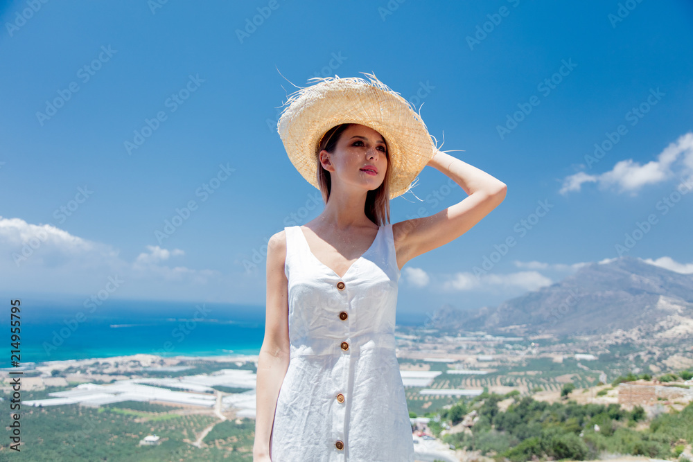 Young woman in white dress and hat is posing on olive garden and sea coast background, Falassandra region, Crete, Greece