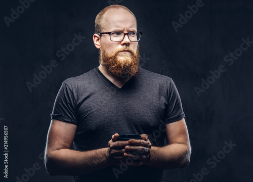 Close up portrait of a redhead bearded male standing with a warming cup of coffee wearing glasses dressed in a gray t-shirt, isolated over dark textured background.