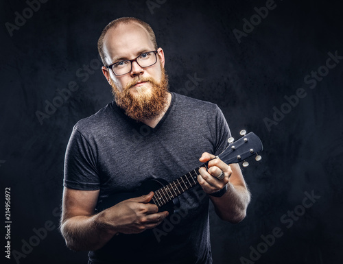 Redhead bearded male musician wearing glasses dressed in a gray t-shirt playing on a ukulele. Isolated on a dark textured background.