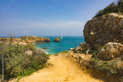 Scenic beach paradise at the Atlantic with the clear blue green water of the ocean and high cliffs with warm colors. The sky is a nice bright blue with white fluffy white clouds.