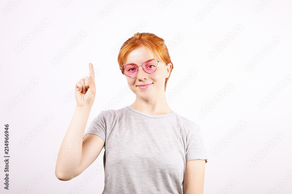 Smiling beautiful young redhead woman pointing to copy space with index finger. Attractive ginger female in hipster pink cat eye glasses showing empty white isolated background. Close up, portrait. Smiling beautiful young redhead woman pointing to copy space with index finger. Attractive ginger female in hipster pink cat eye glasses showing empty white isolated background. Close up, portrait.
