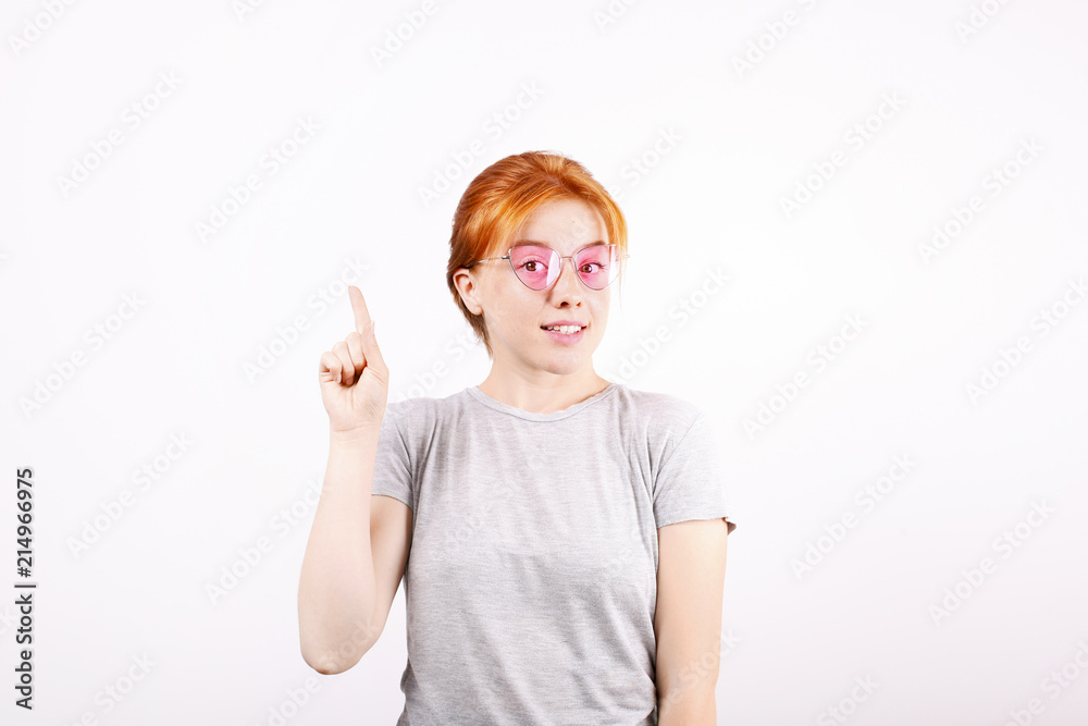 Smiling beautiful young redhead woman pointing to copy space with index finger. Attractive ginger female in hipster pink cat eye glasses showing empty white isolated background. Close up, portrait. Smiling beautiful young redhead woman pointing to copy space with index finger. Attractive ginger female in hipster pink cat eye glasses showing empty white isolated background. Close up, portrait.