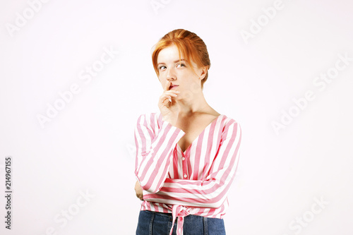 Young beautiful woman with long natural red hair, thinking facial expression, hands on chin thinker pose. Attractive thoughtful hipster redhead female in pink white knot shirt. Background, copy space.