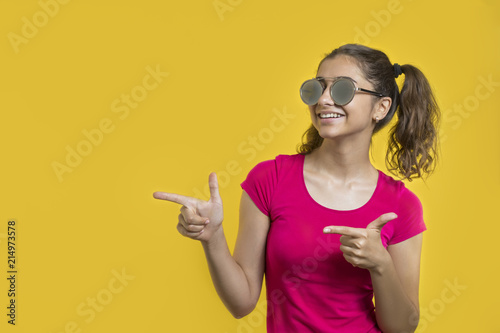 Happy, smiling girl in sunglasses on yellow background points with fingers on copy space. 
