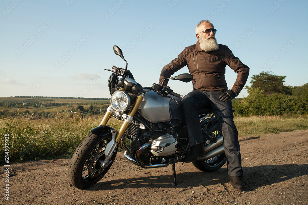 Old rider biker man in black leather jacket jeans boots and helmet sit on classic style cafe racer motorcycle. Brutal fun urban lifestyle. Outdoor portrait. Stock Adobe Stock