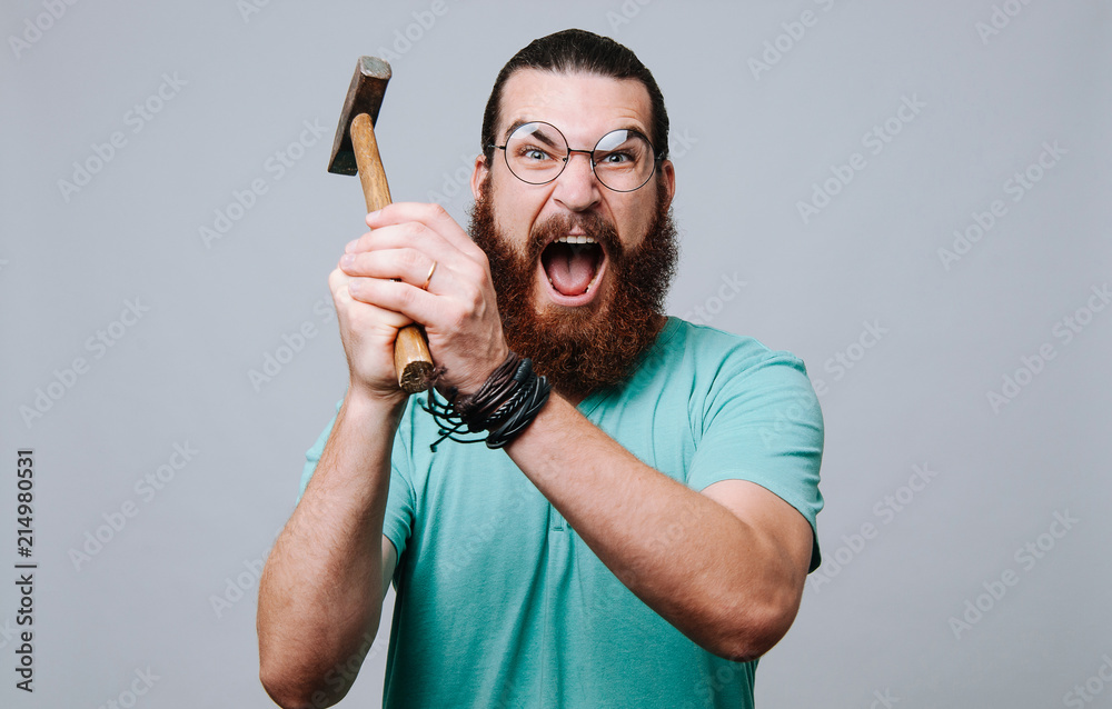 Bearded young man looking and screaming at the camera holding a hammer and ready to hit. - obrazy, fototapety, plakaty 