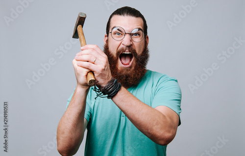 Slika na platnu Bearded young man looking and screaming at the camera holding a hammer and ready to hit