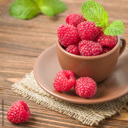 Ripe fresh raspberries with green mint leaves in brown cup and saucer on wooden background.