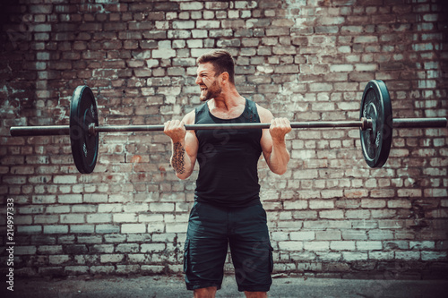 Athletic man working out with a barbell in front of brick wall. Strength and motivation. Outdoor workout. Biceps exercise. photo