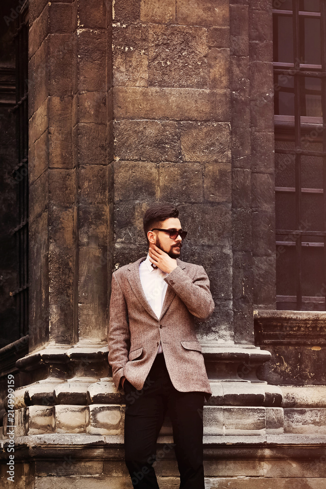 Young man in stylish suit in the old town. The guy poses on the camera and touches his beard. Italian look Young man in stylish suit in the old town. The guy poses on the camera and touches his beard. Italian look