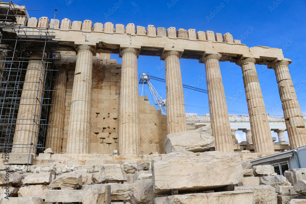 Looking up at the Athens Parthnon being reconstructed with pieces numbered and placed on wooden stands in the foreground and a tall crane and very blue sky seen through the rebuilt columns