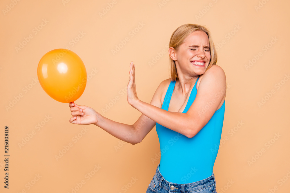 Portrait of young attractive straight-haired blonde girl wearing casual, jeans shorts. Keeping yellow air baloon, scared of it. Isolated over beige background