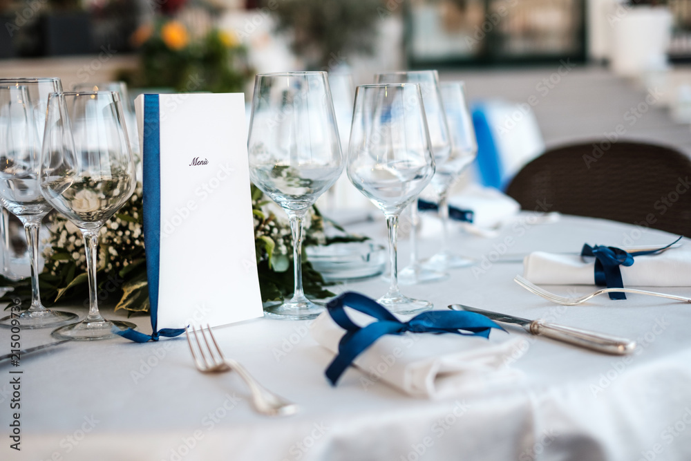 Prospettiva di una tavola imbandita con bicchieri di vino e posate per una  cerimonia in un ristorante italiano, con posate, fiocchi blu ed un menù in  evidenza. Stock Photo | Adobe Stock