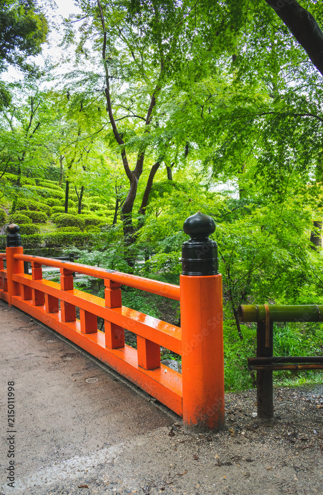 A partial view of an orange painted wooden bridge in the park or garden of Fushimi inari Teisha shrine in Kyoto, Japan, during a summer day in rainy season A partial view of an orange painted wooden bridge in the park or garden of Fushimi inari Teisha shrine in Kyoto, Japan, during a summer day in rainy season