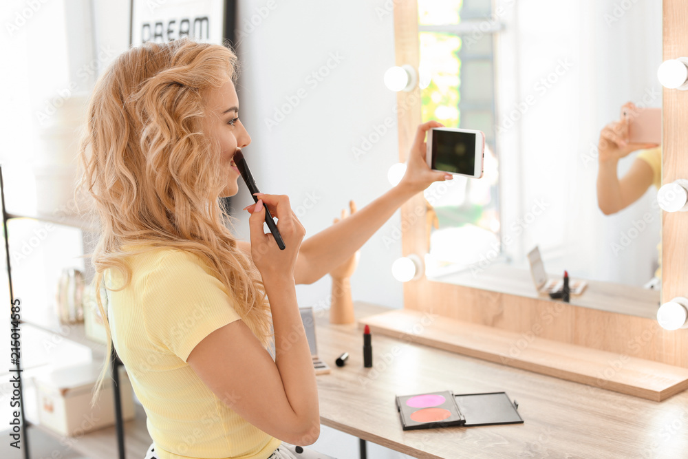 Beauty blogger filming makeup tutorial with smartphone in front of mirror at dressing table indoors - obrazy, fototapety, plakaty 