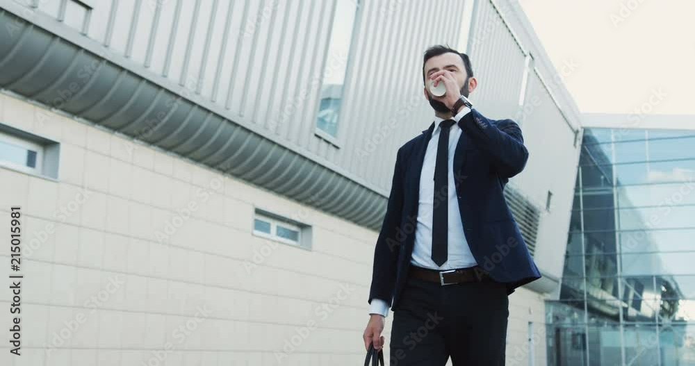 Caucasian office worker in a suit and tie with a bag drinking coffee in the morning before work near office building. Young businessman going to meeting. Outdoor. Caucasian office worker in a suit and tie with a bag drinking coffee in the morning before work near office building. Young businessman going to meeting. Outdoor.