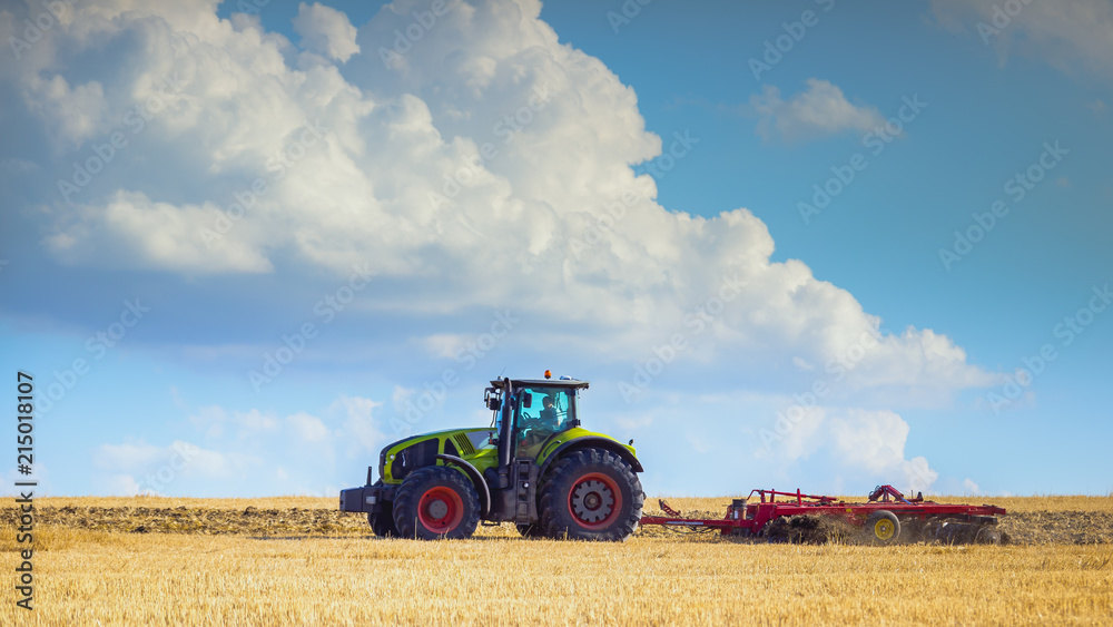 Obraz premium In the summer heat of the tractor plows the field after harvest against the bright Sunny sky with clouds.