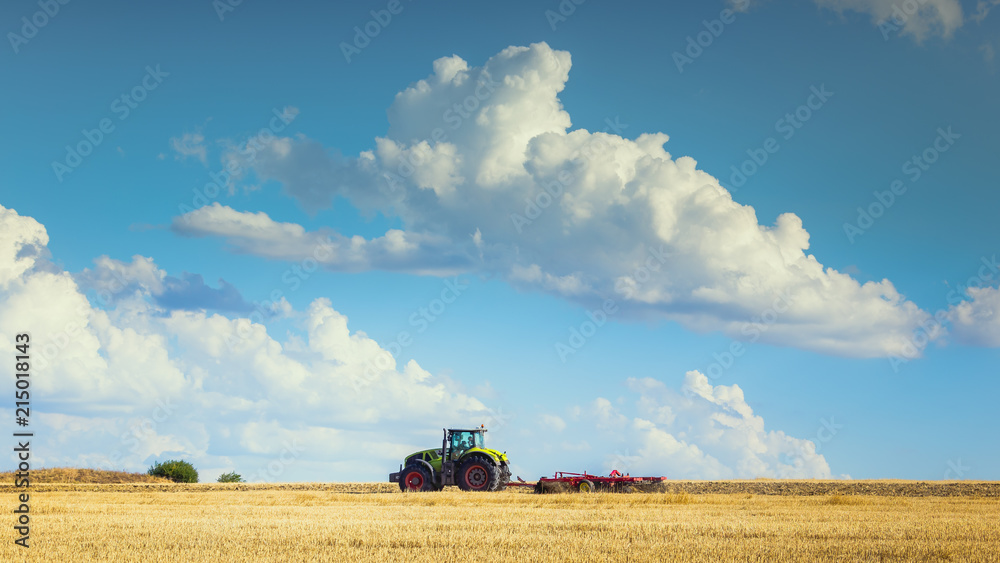 Fototapeta premium In the summer heat of the tractor plows the field after harvest against the bright Sunny sky with clouds.