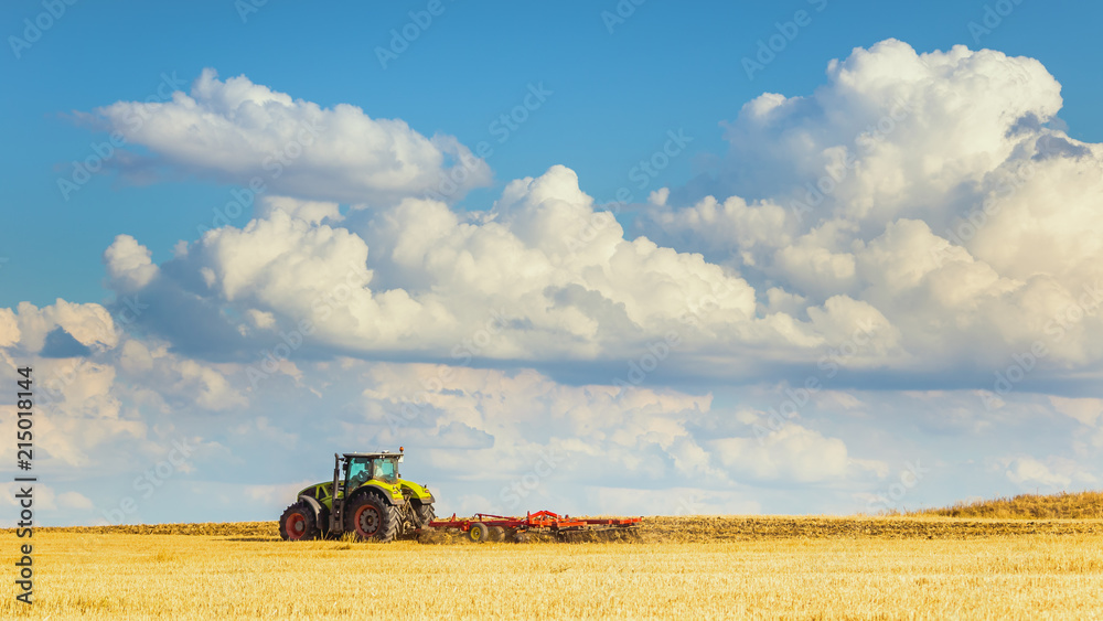 Fototapeta premium In the summer heat of the tractor plows the field after harvest against the bright Sunny sky with clouds.