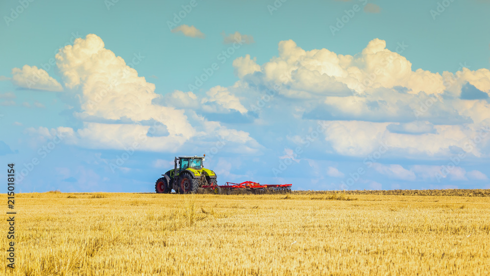 Fototapeta premium In the summer heat of the tractor plows the field after harvest against the bright Sunny sky with clouds.