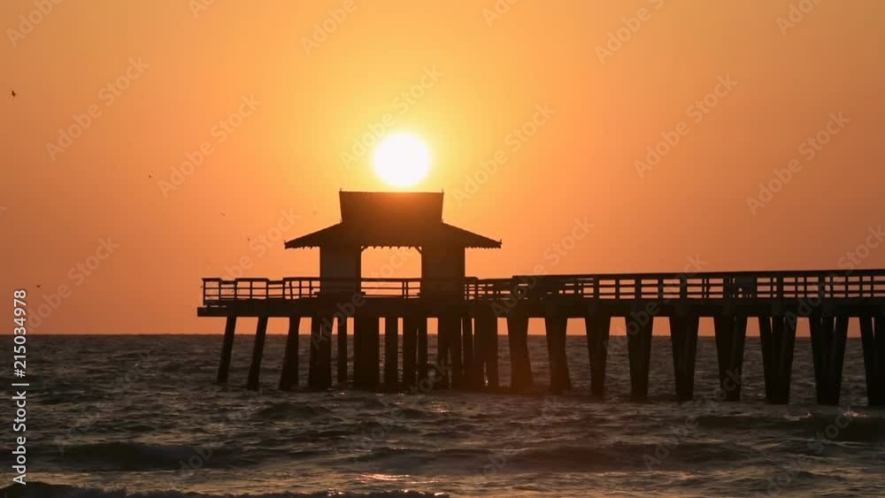 Naples, Florida yellow and orange sunset in gulf of Mexico with sun setting inside Pier wooden jetty, with many birds silhouette flying over horizon and dark blue ocean waves Naples, Florida yellow and orange sunset in gulf of Mexico with sun setting inside Pier wooden jetty, with many birds silhouette flying over horizon and dark blue ocean waves
