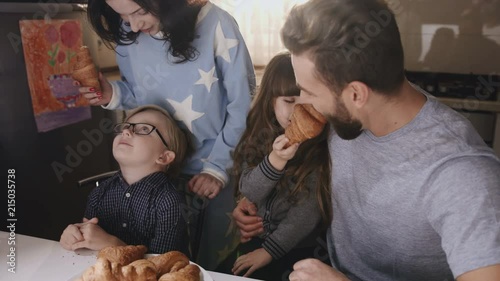 Happy family of parents and two little children sitting at the table in the kitchen in the morning and eating croissants. Portrait shot. Indoor photo