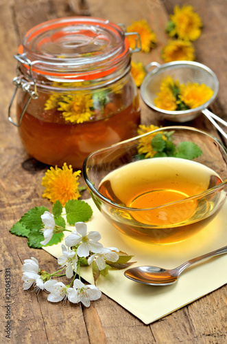 Dandelion honey, spring flowers, spoon, dandelion head around, small colander and full jar in background - vertical photo
