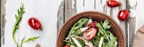 Closeup of delicious salad of arugula with cherry tomatoes and chicken breast in a clay bowl on a wooden table, top view. Sports nutrition. Balanced meals.