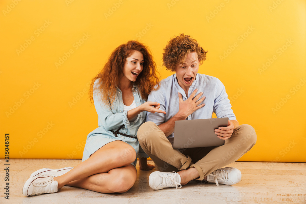 Photo of amazed beautiful couple man and woman 20s sitting on floor and using silver laptop, isolated over yellow background Photo of amazed beautiful couple man and woman 20s sitting on floor and using silver laptop, isolated over yellow background
