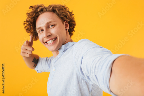 Photo of young beautiful man 20s taking selfie photo and pointing finger at camera, isolated over yellow background
