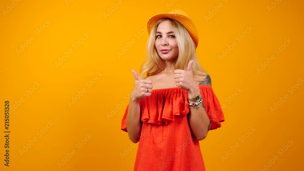 Blonde sexy woman in summer dress shows thumbs up to the camera isolated on orange yellow background. Successful woman showing gestures. Copyspace available Blonde sexy woman in summer dress shows thumbs up to the camera isolated on orange yellow background. Successful woman showing gestures. Copyspace available