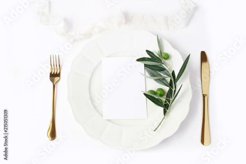 Festive table summer setting with golden cutlery, olive branch, porcelain dinner plate and silk ribbon on white table background. Blank card mockup. Mediterranean wedding or restaurant menu concept