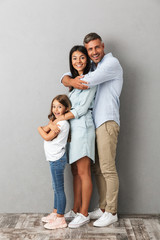 Full length photo of beautiful caucasian family woman and man with little girl smiling and hugging together, isolated over gray background