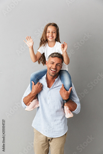 Photo of joyful family smiling at camera while little girl having fun and sitting on the neck of her happy father, isolated over gray background