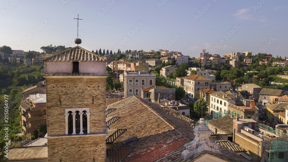 Aerial detail of a bell tower of the Castelnuovo church with a cross on the summit. Behind the tower opens a valley with green trees and plants. You can see the roofs of the surrounding buildings.